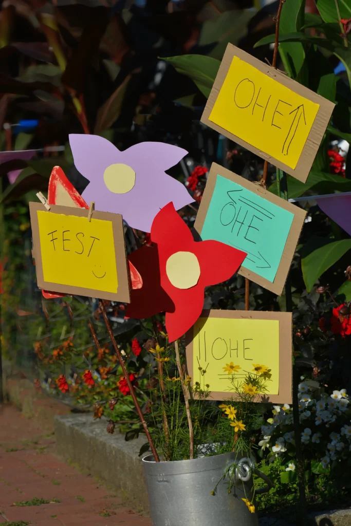 Gebastelte bunte Papierblumen und Pappschilder mit dem Wort Ohe, an Holzstangen, die im Blumenbeet stecken.